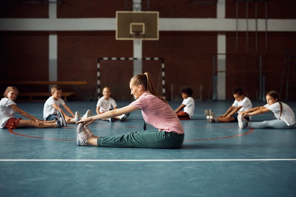 Happy coach and group of elementary students doing stretching exercises on PE class at school gym.